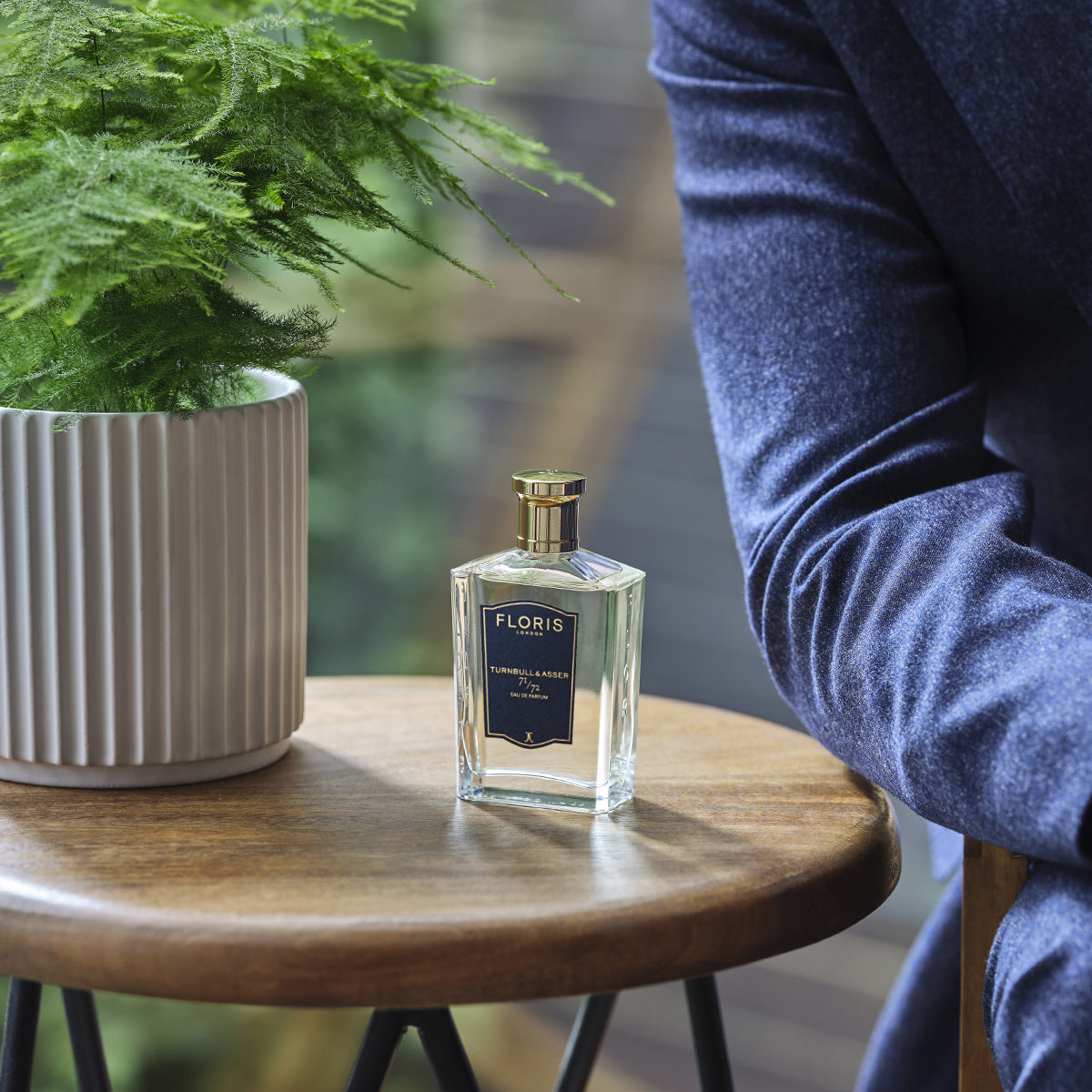 A bottle of Floris London 71/72 Eau de Parfum rests on a wooden table by a potted plant, next to a blue velvet men’s jacket.