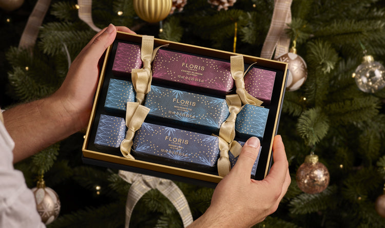 Three beautifully decorated crackers displayed in a gift box. The box is being held in front of a decorated Christmas tree.