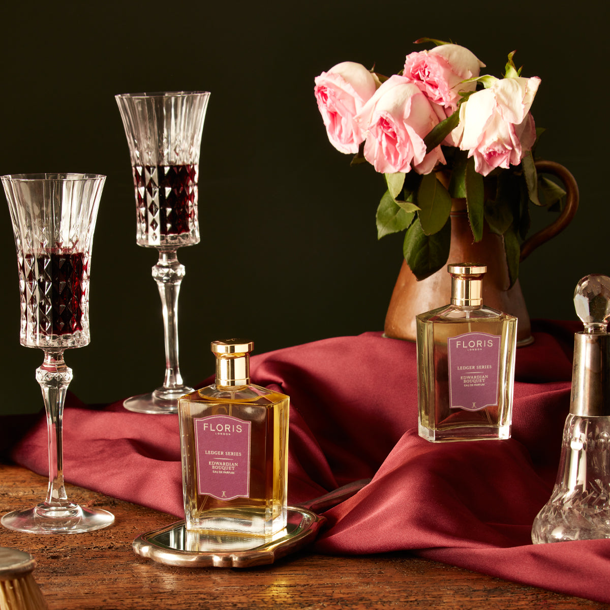 A still life with two wine glasses, two Edwardian Bouquet perfumes by Floris London EU, pink roses in a ceramic jug, a salt shaker, and a red cloth on wood.