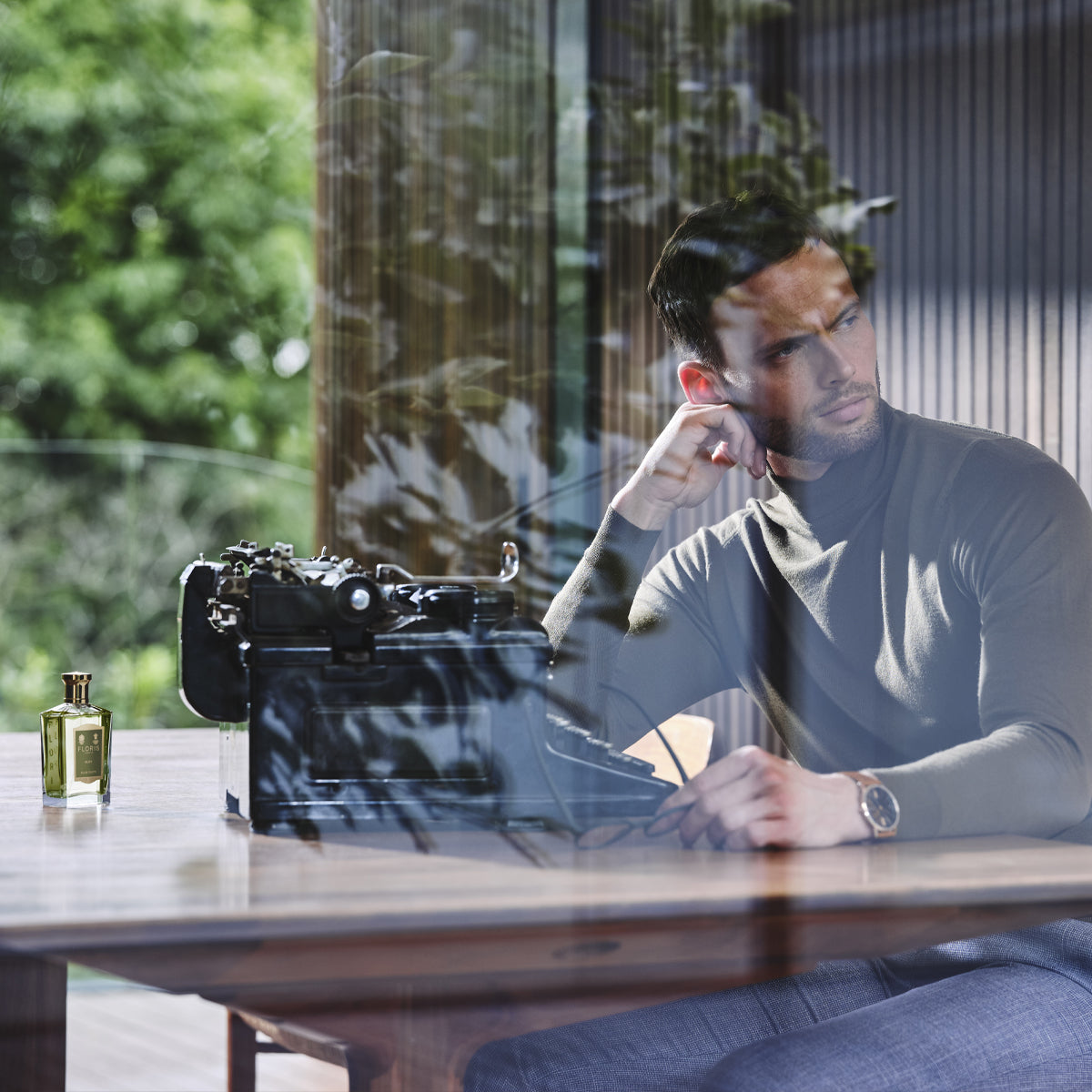 A man in a turtleneck sits at a wooden desk with a vintage typewriter and Floris London Elite Eau de Toilette, gazing thoughtfully out the window.