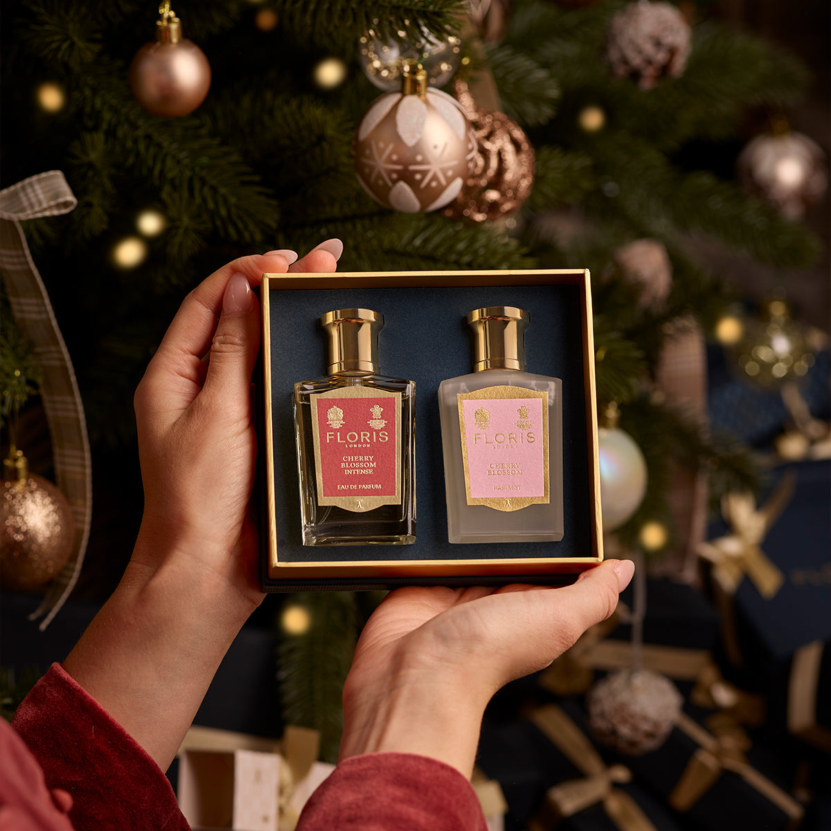 Hands holding a Cherry Blossom Duet gift set from Floris London EU, featuring two perfume bottles, with a decorated Christmas tree and wrapped presents in the background, evoking an air of festive intensity.