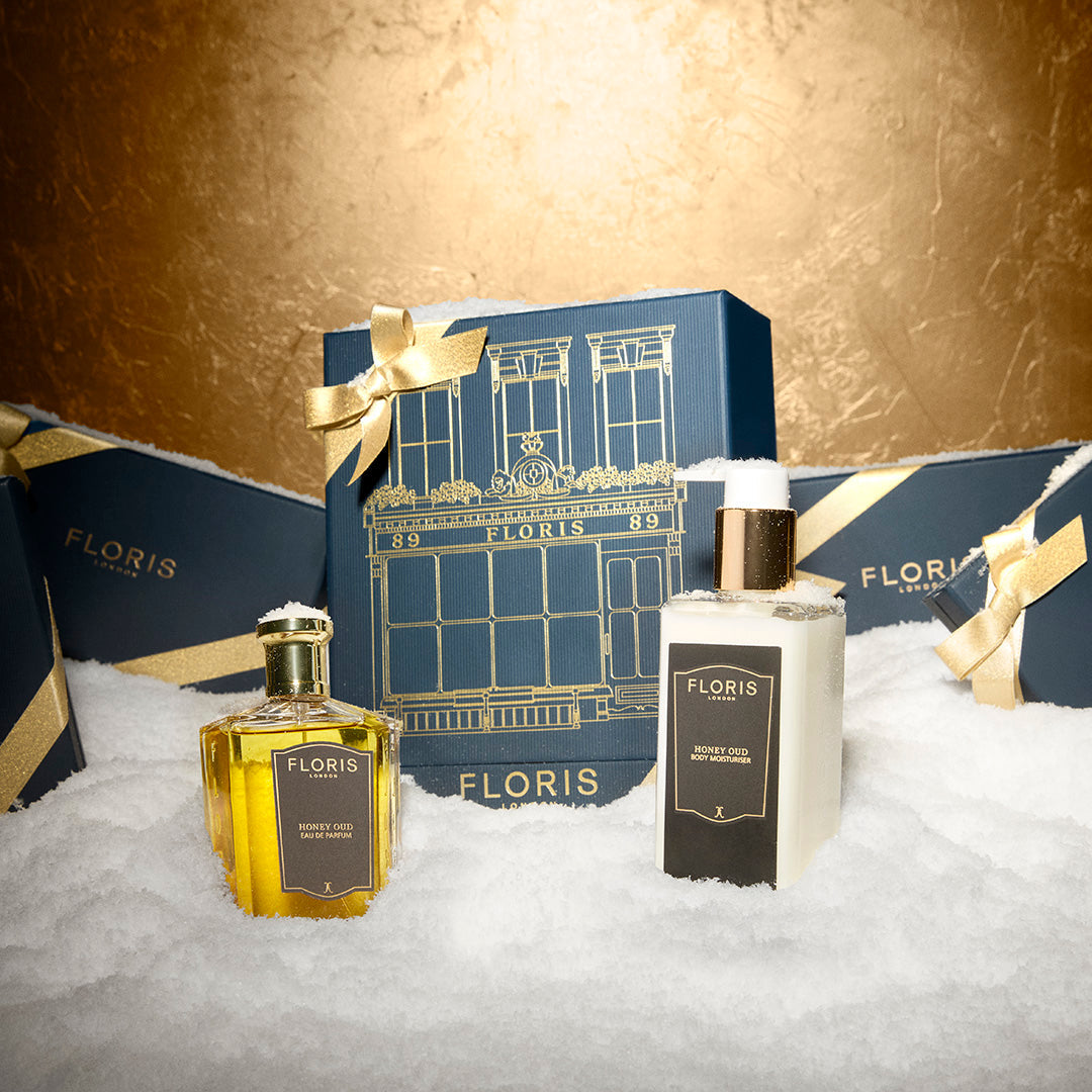 Two Floris London EU Sweet Lux | Honey Oud Duo bottles sit in snow with a blue gift box tied in gold ribbon, plus more gift boxes behind them.
