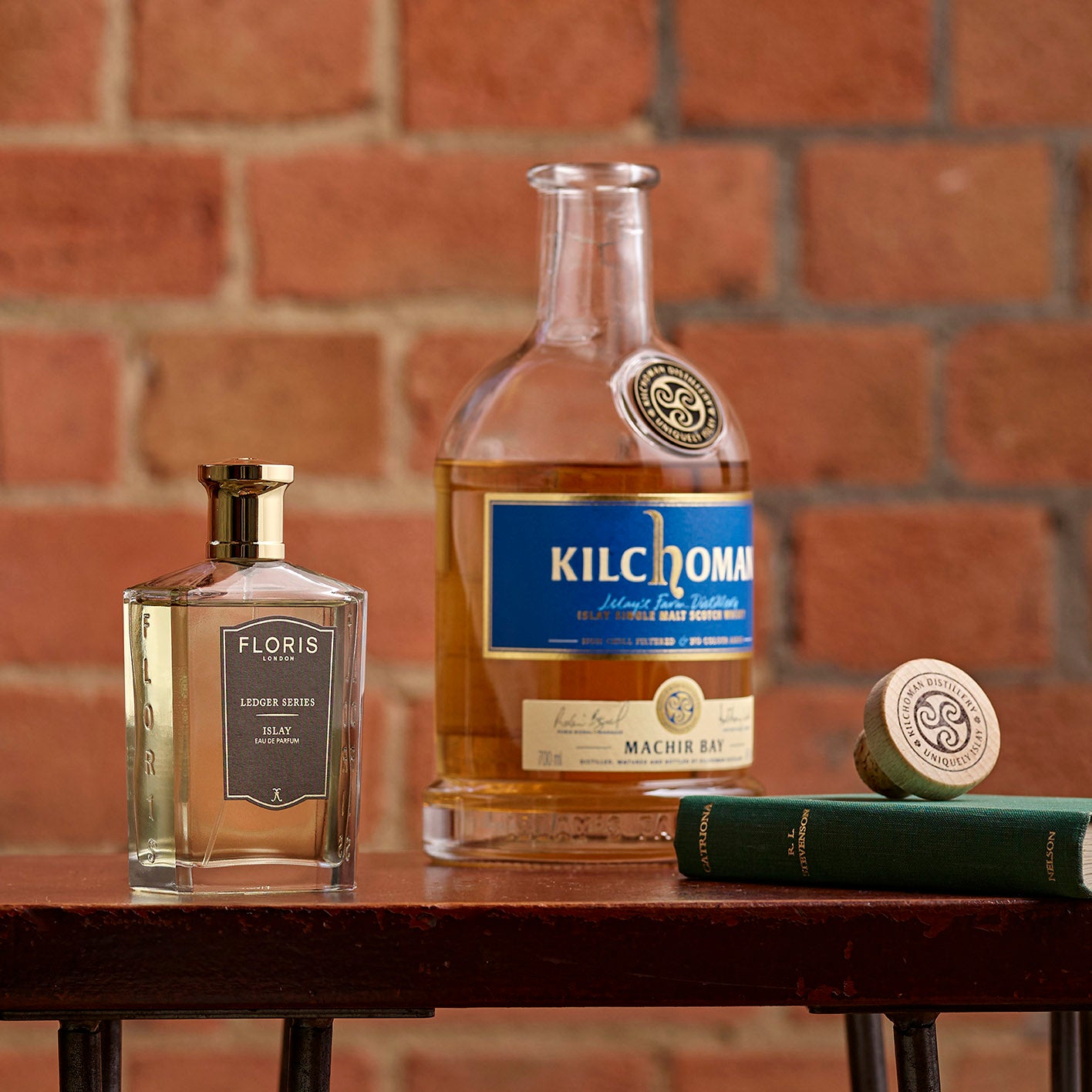 A bottle of Floris London EU's Islay fragrance rests on wood, with a book and rustic brick wall creating a warm, inviting setting.