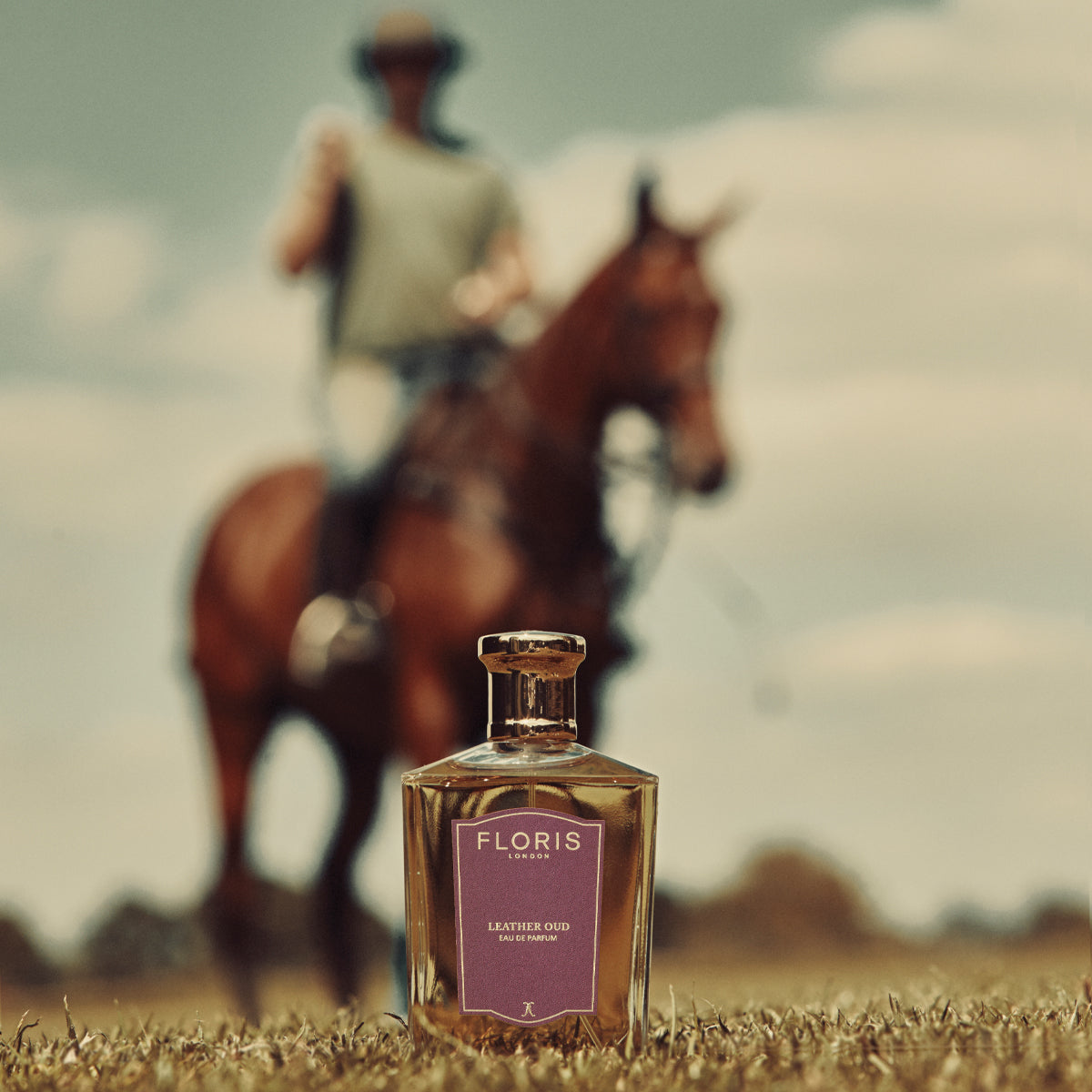 A bottle of Floris London Leather Oud - Eau de Parfum lies on grass while a blurred horseback rider passes in the background.