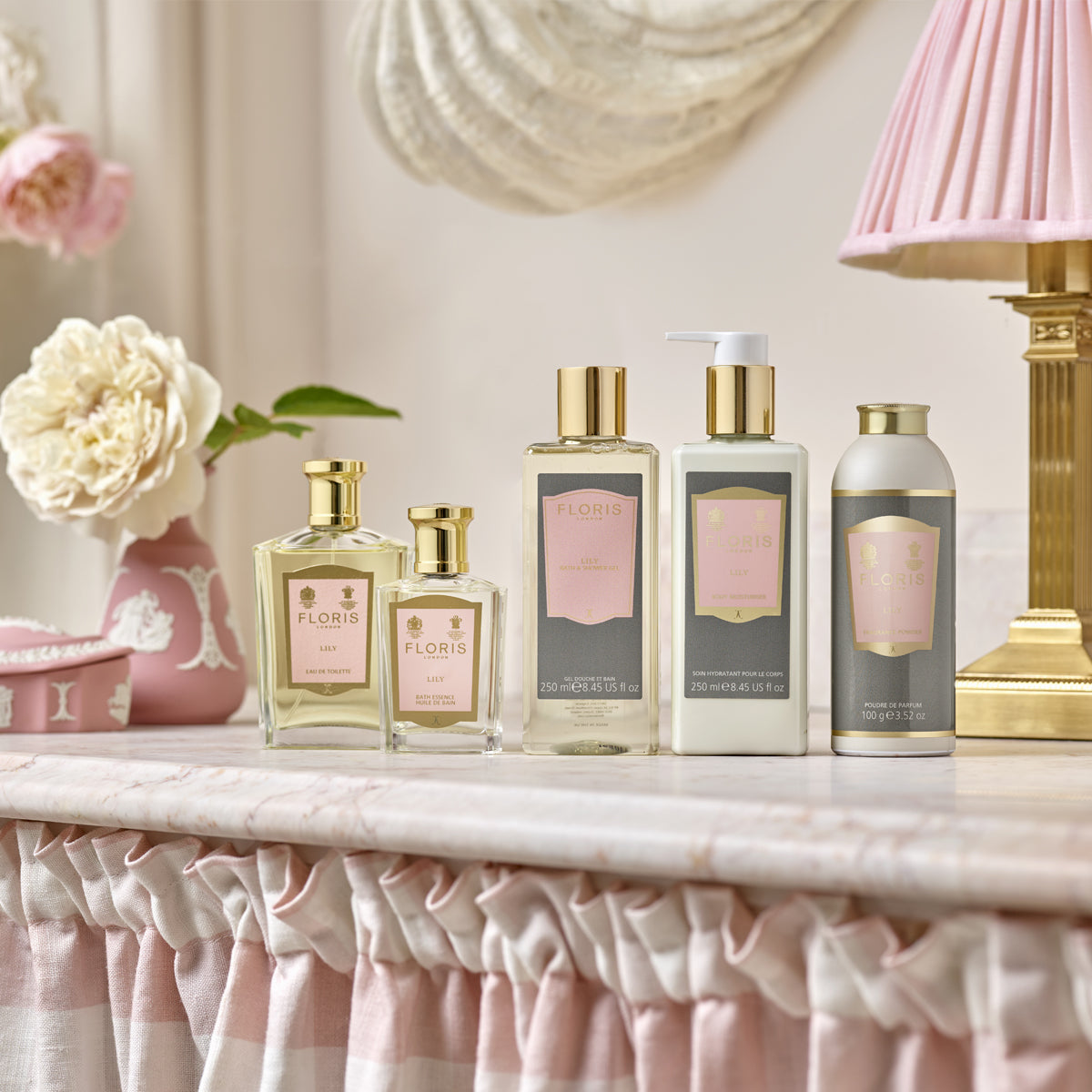Five Floris London EU Lily - Eau de Toilette toiletries with a floral scent sit on a marble counter with a pink ruffled cloth, lamp, and flowers.