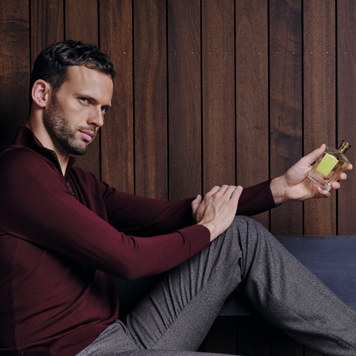A man in a burgundy sweater and gray pants sits by a wooden wall, thoughtfully holding Floris London Limes - Eau de Toilette.