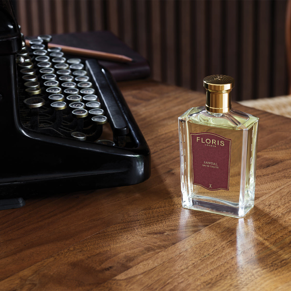 A bottle of Floris London Santal Eau de Toilette, a woody amber men’s fragrance, sits on a wooden desk beside a vintage typewriter and closed notebook.