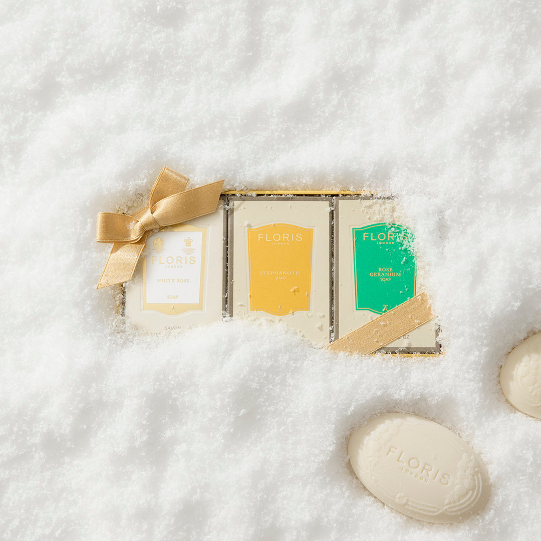 A gold gift box, Soap Collection | For Her by Floris London EU, with three soaps—two tied in gold ribbon—rests in snow beside two loose bars.