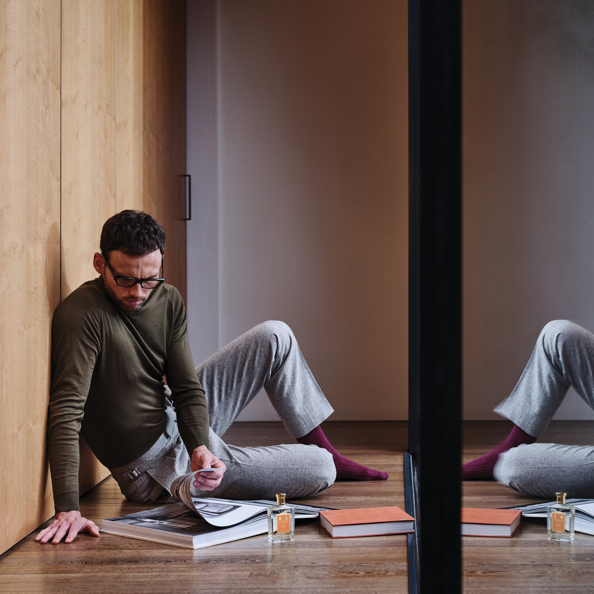 A man in glasses sits by a wardrobe, reading a magazine, with books and a bottle of Floris London Special No. 127 Eau de Toilette reflected in glass.