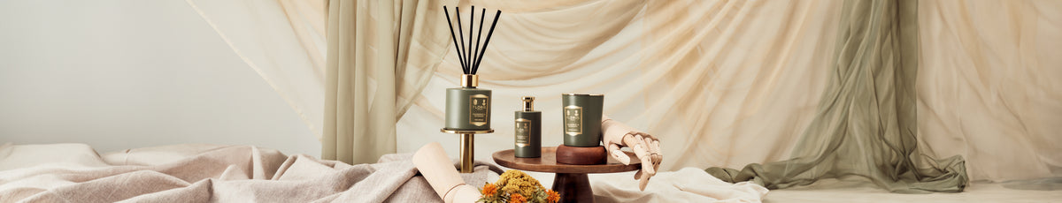 Elegant green and gold diffusers and lotions on a wooden tray with draped fabric backdrop.