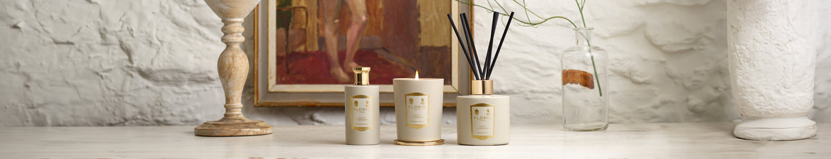 Candles and a reed diffuser on a table with a rustic white wall and framed artwork in the background.