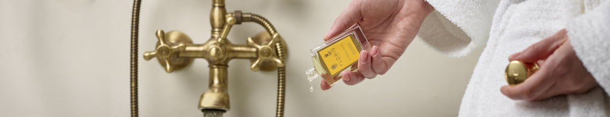 A person in a bathrobe holding a small bottle of yellow liquid near a vintage brass faucet.