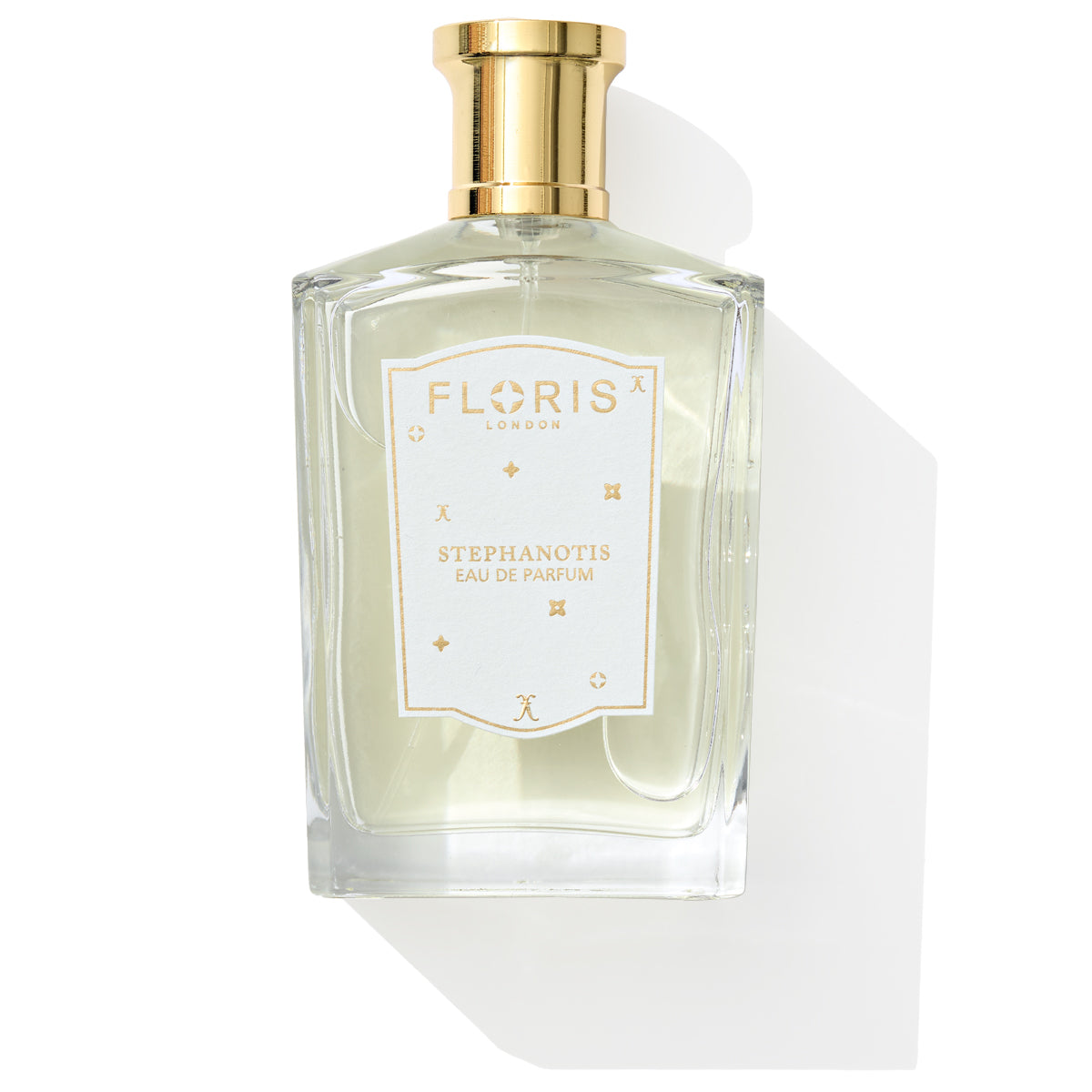 A clear glass bottle of Stephanotis - Eau de Parfum | Limited Edition by Floris London EU, featuring a gold cap and white label, cast a shadow on a pristine white background.