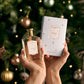Hands holding a bottle of Stephanotis - Eau de Parfum | Limited Edition by Floris London EU and its matching box, set against a background featuring a decorated Christmas tree.
