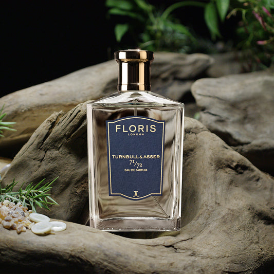 A glass bottle of Floris London 71/72 - Eau de Parfum, a Jermyn Street scent for men, sits on driftwood amid small plants and stones.