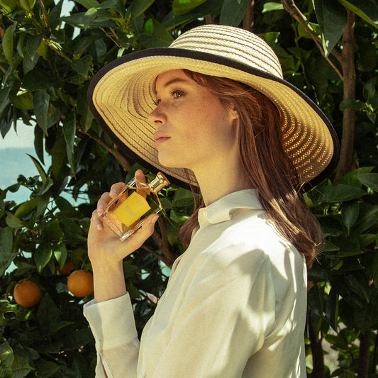 A woman in a wide-brimmed hat and white shirt holds Floris London's Bergamotto di Positano Eau de Parfum amid green foliage and orange trees.