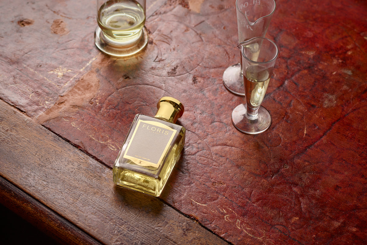 A 50ml Floris London Customisation Replenishment bottle with a gold cap and label, resting on worn leather beside two glass beakers.