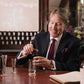 A man in a suit smiles as he pours Floris London’s Fragrance - Customisation Replenishment 100ml into a glass, with bottles and a notebook nearby.
