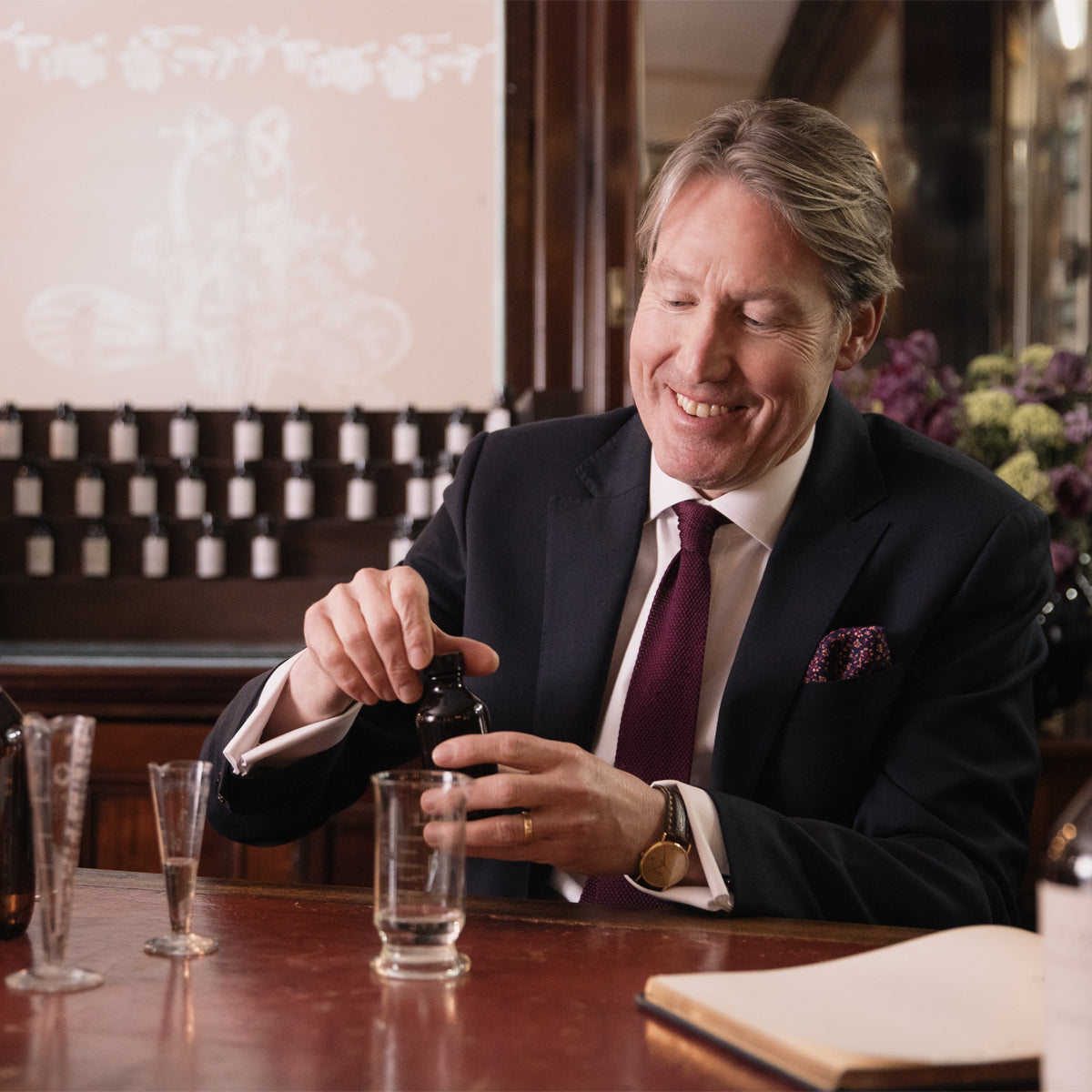 A man in a suit smiles as he pours Floris London’s Fragrance - Customisation Replenishment 100ml into a glass, with bottles and a notebook nearby.