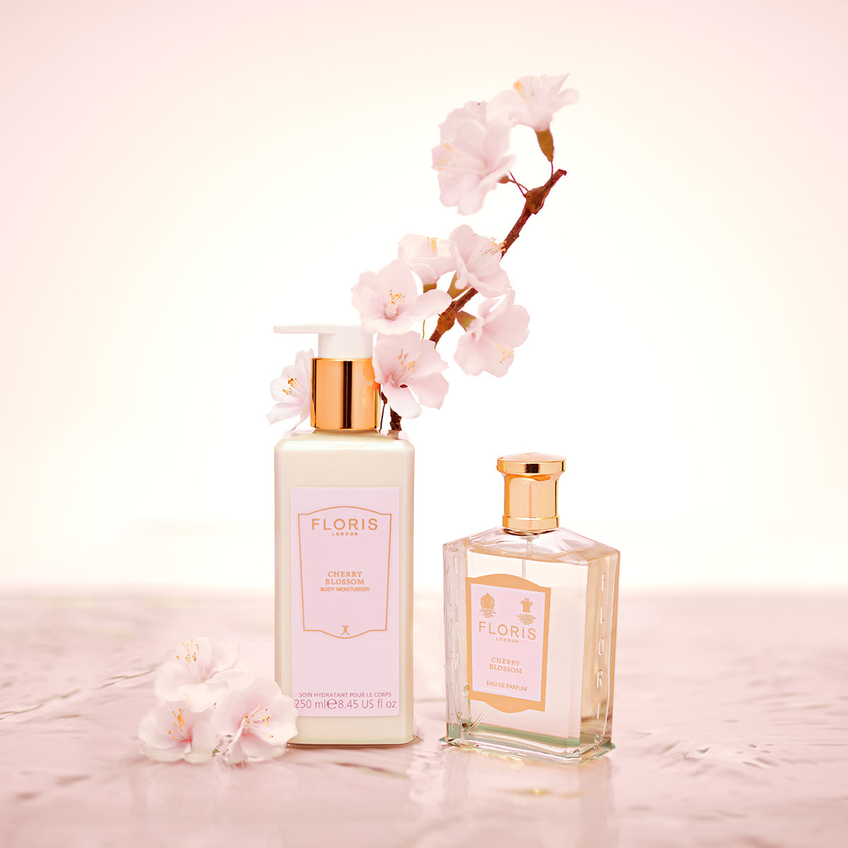 Two Floris London UK Cherry Blossom - Enriched Body Moisturiser bottles with gold caps beside pink cherry blossoms on a pink surface.