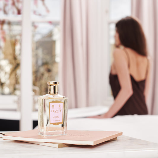 A bottle of Floris London Cherry Blossom - Eau de Parfum rests on a pink book, its floral-fruity scent evoking spring, woman in brown blurred behind.