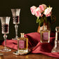 A still life with two wine glasses, two Edwardian Bouquet perfumes by Floris London EU, pink roses in a ceramic jug, a salt shaker, and a red cloth on wood.
