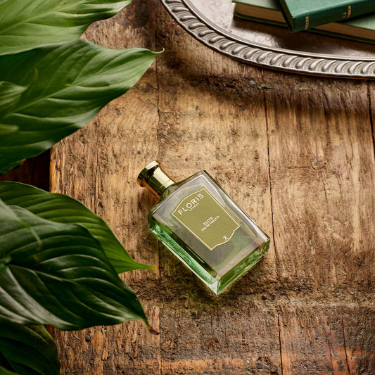 A bottle of Elite - Eau de Toilette by Floris London sits on rustic wood, surrounded by green leaves, with books and a silver tray nearby.