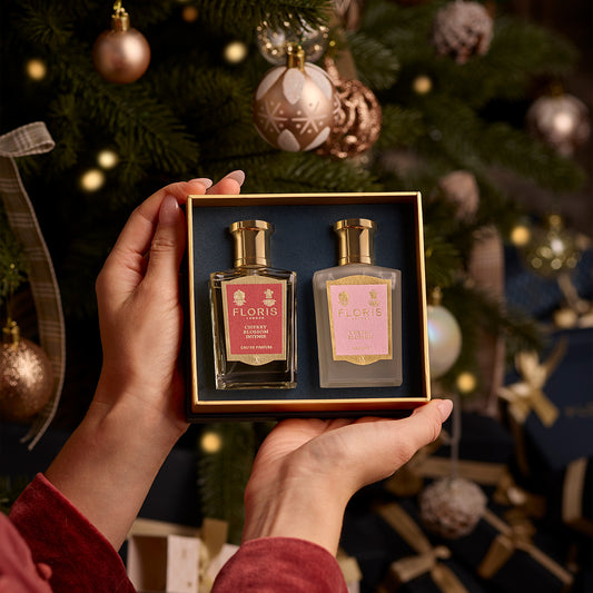 Hands holding a Cherry Blossom Duet gift set from Floris London EU, featuring two perfume bottles, with a decorated Christmas tree and wrapped presents in the background, evoking an air of festive intensity.
