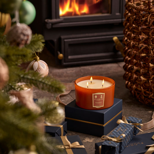 A lit Cinnamon & Tangerine Deluxe Three Wick Candle, a limited-edition product from Floris London EU, sits on a box in front of a fireplace with wrapped presents and a decorated Christmas tree nearby in a cozy indoor setting.