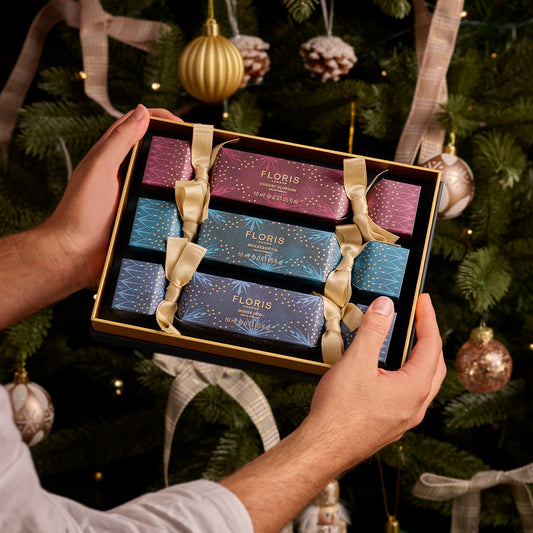 Hands holding a box of the Holiday Cracker Collection by Floris London EU, featuring three festive holiday crackers tied with gold ribbons, in front of a decorated Christmas tree—all part of a cherished British tradition during the holiday season.