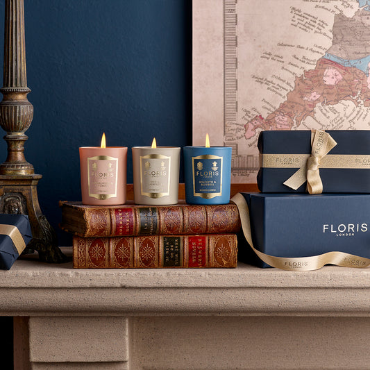 Three beautifully scented candles from the Miniature Candle Collection by Floris London EU are placed on antique books, next to a ribbon-adorned box and a lamp. A decorative map is visible in the background.