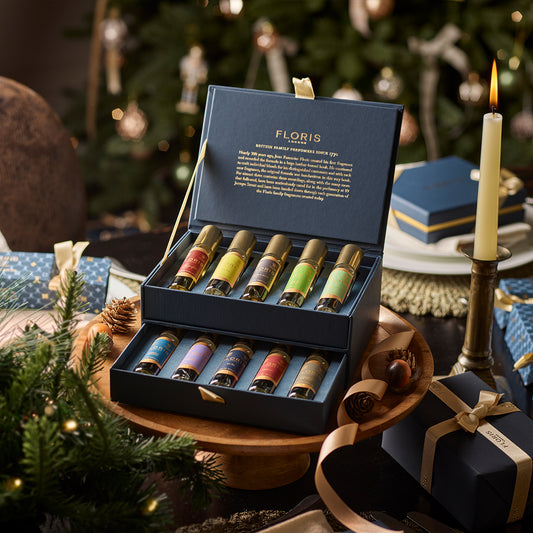 A box of assorted perfumes from The Perfumer's Collection by Floris London, with festive decor in the background, featuring a lit candle and wrapped gifts, makes for the perfect gift for any fragrance connoisseur.