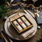 A dinner setting features a square gift box of Floris London EU's "Perfumer's Trilogy: For Him" on a plate, with silverware and ribbon decorations completing the scene.