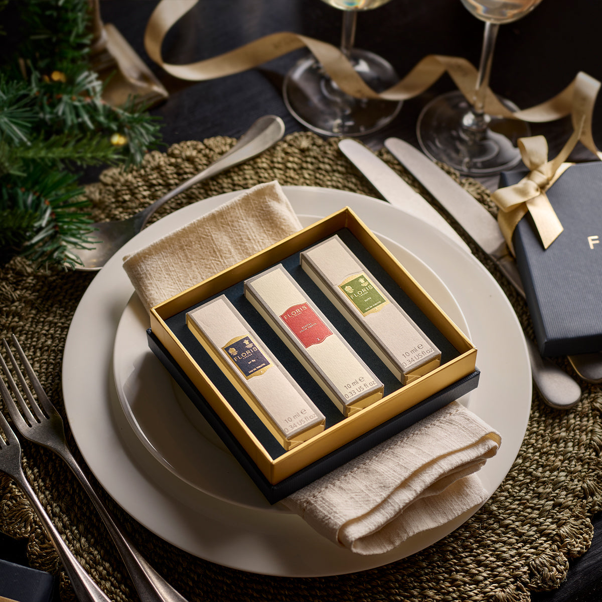 A dinner setting features a square gift box of Floris London EU's "Perfumer's Trilogy: For Him" on a plate, with silverware and ribbon decorations completing the scene.