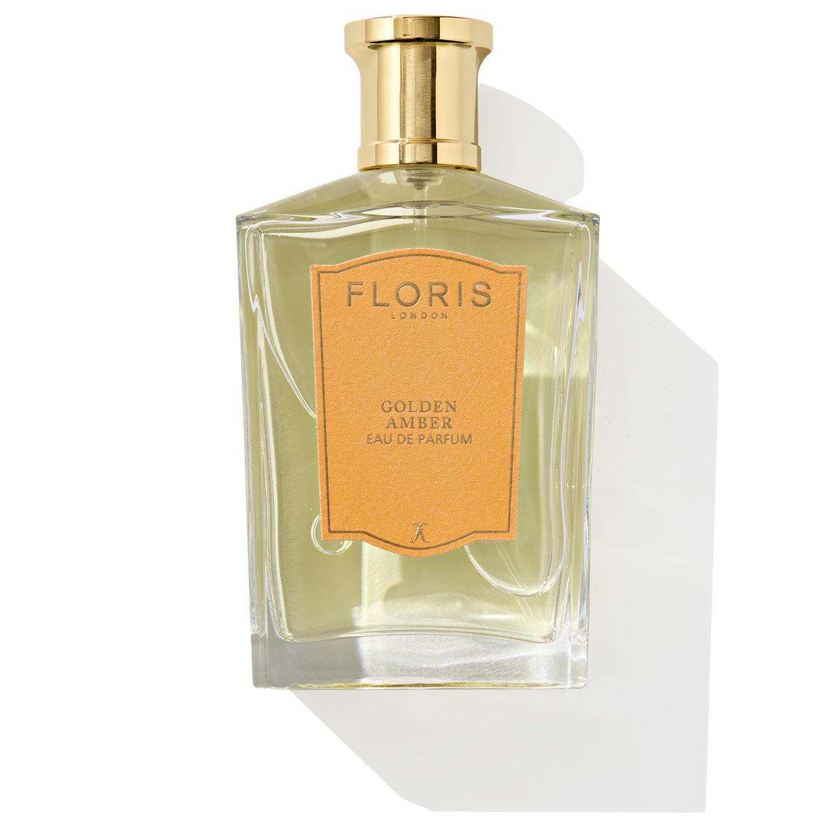 A bottle of Floris London's Golden Amber - Eau de Parfum, featuring a gold cap and an amber-colored label, casts a shadow to the right. The fragrance combines a floral heart with subtle aquatic notes, resulting in an elegant and refreshing scent.