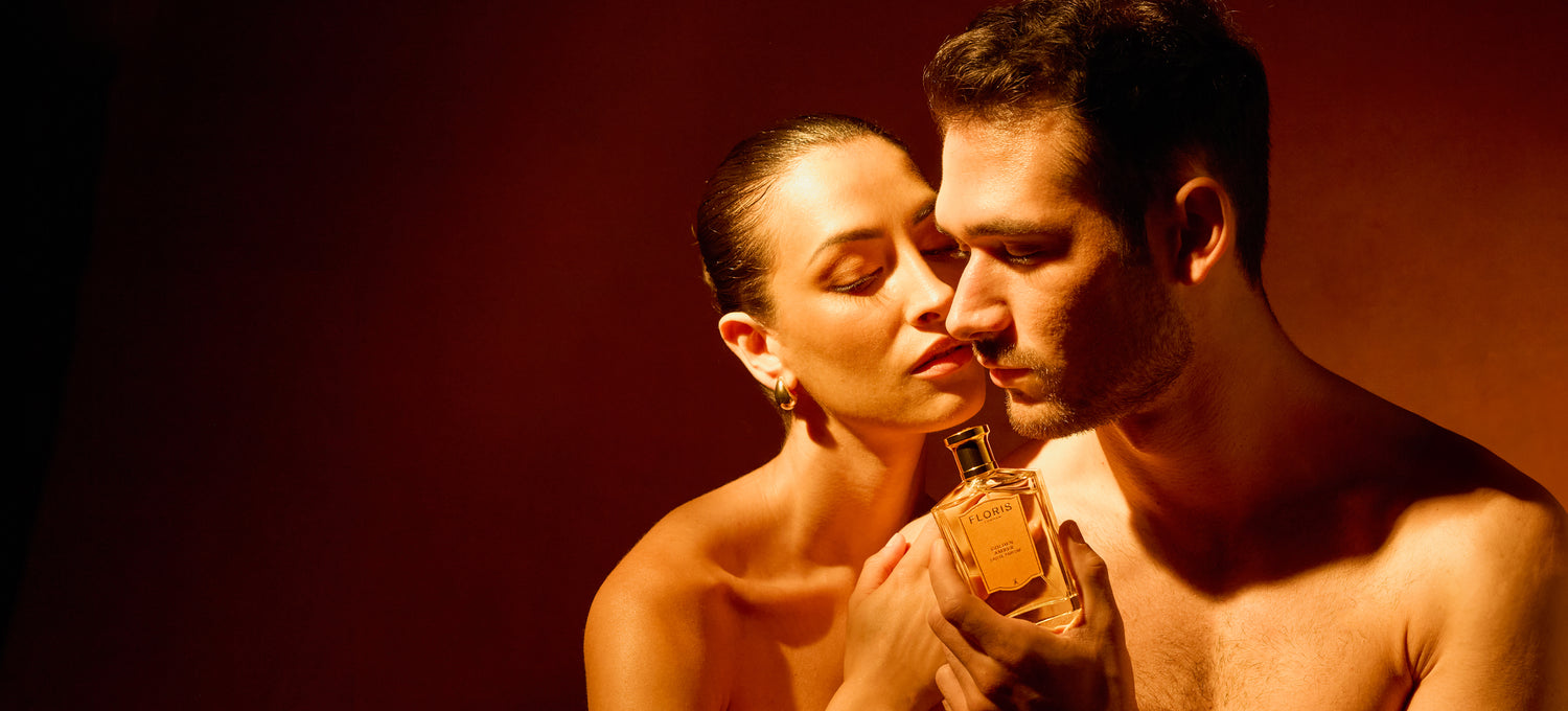 A woman and a man pose closely, with the man holding a bottle of Floris perfume against a dark, warm-toned background.