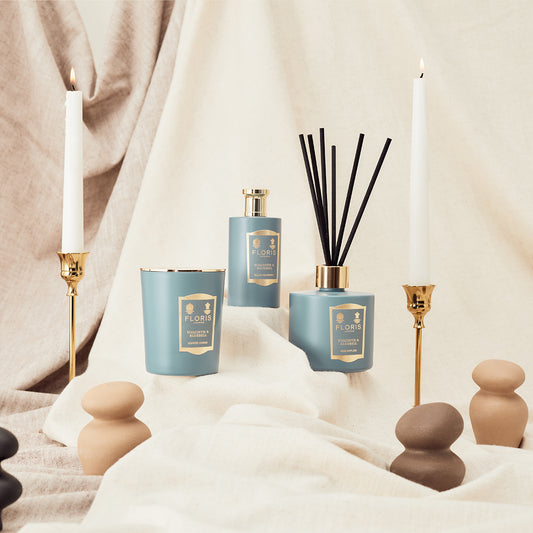 Floris London’s Hyacinth & Bluebell Reed Diffuser, white candles in gold holders, and smooth sculptures are arranged on draped beige fabric.