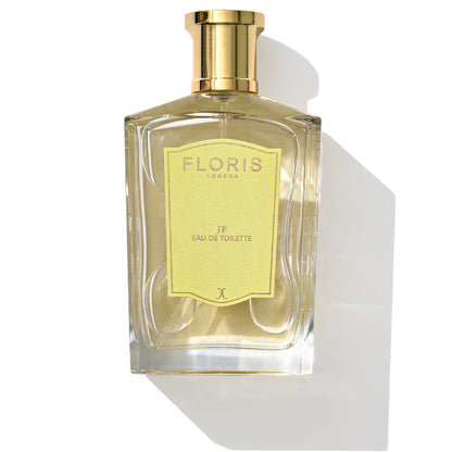 A clear glass bottle with a gold-colored cap containing JF - Eau de Toilette by Floris London, a signature fragrance for men. The label is yellow with gray text. The bottle casts a shadow, echoing its citrus and woody scent.