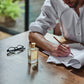 A person in a white shirt writes on papers at a wooden table with glasses, beside Floris London’s JF - Eau de Toilette men’s perfume.