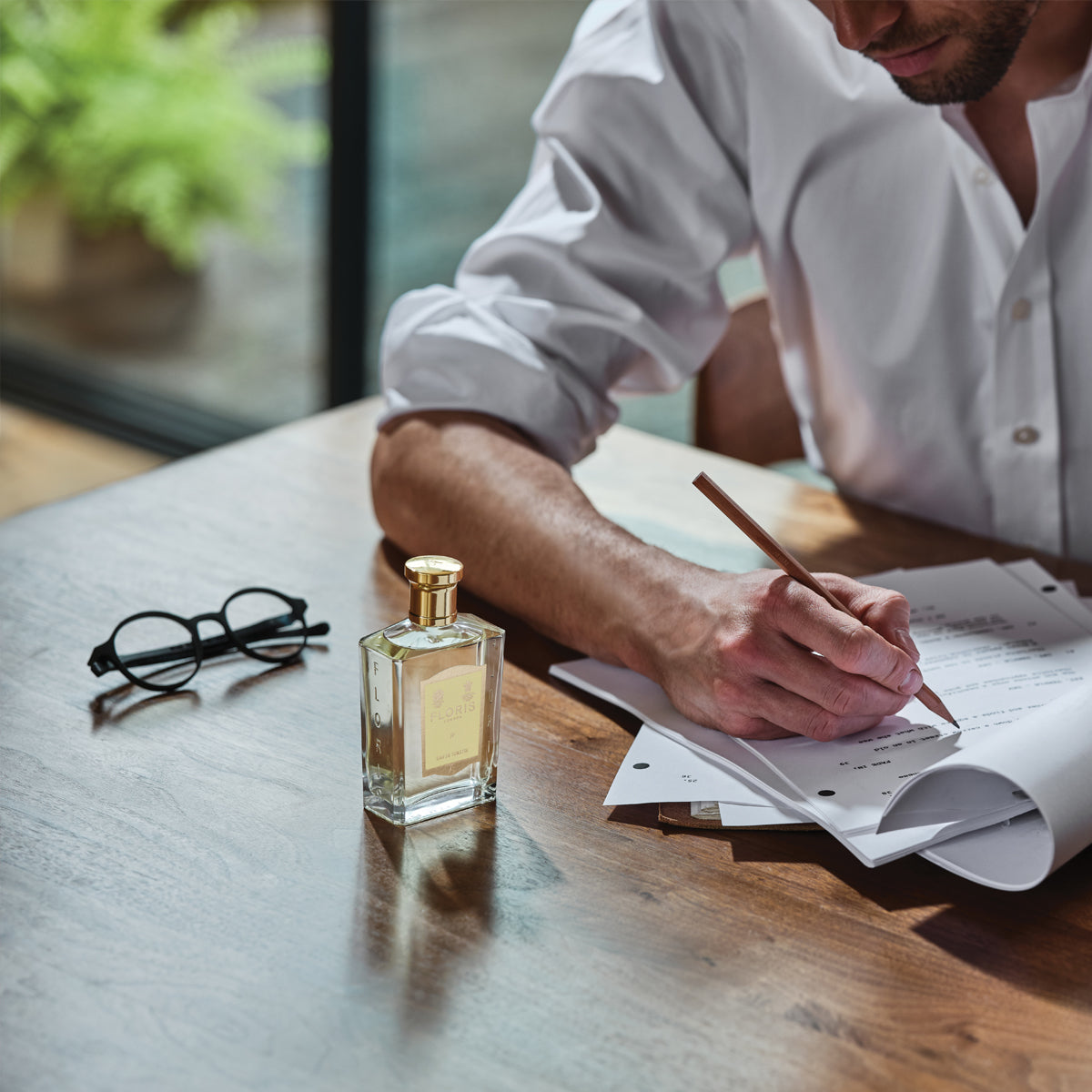 A person in a white shirt writes on papers at a wooden table with glasses, beside Floris London’s JF - Eau de Toilette men’s perfume.
