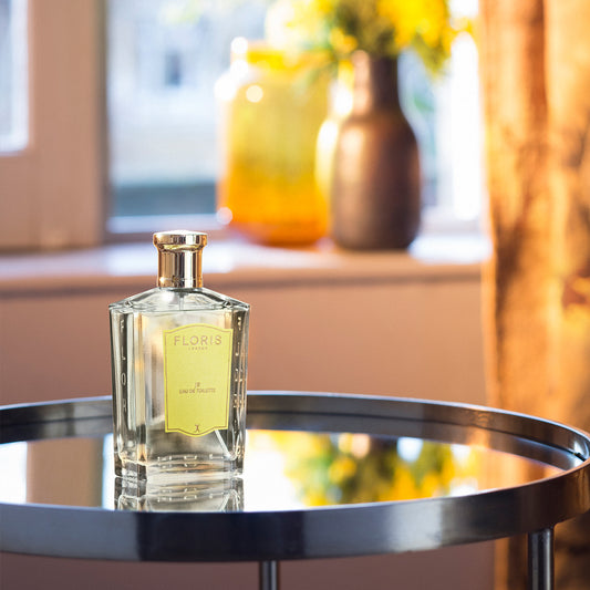 A bottle of Floris London JF - Eau de Toilette rests on a round glass table, with blurred vases and a window in the background.