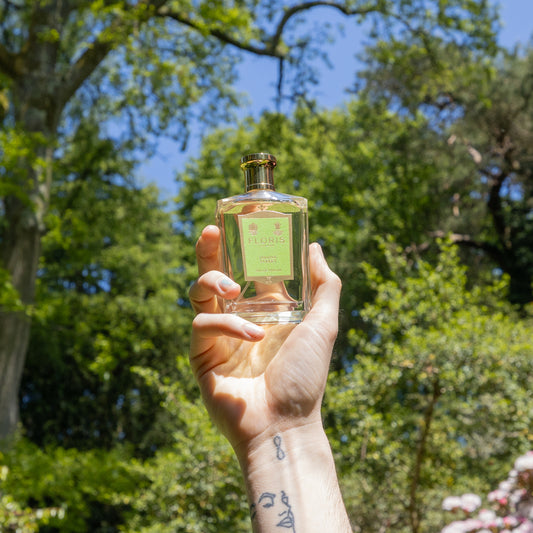 A hand with a line-art face tattoo holds up a clear Jermyn Street Eau de Parfum bottle by Floris London outdoors, with trees and sky behind.