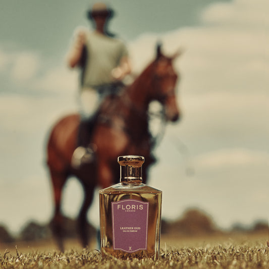 A bottle of Floris London Leather Oud - Eau de Parfum lies on grass while a blurred horseback rider passes in the background.