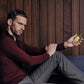 A man in a burgundy sweater and gray pants sits by a wooden wall, thoughtfully holding Floris London Limes - Eau de Toilette.