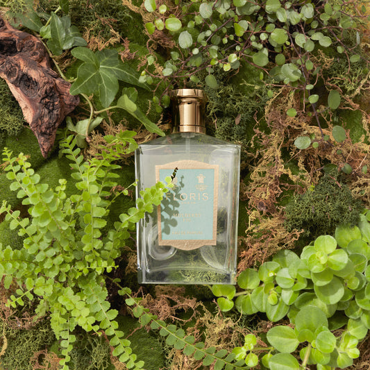 The Mulberry Fig Eau de Parfum by Floris London, in a rectangular glass bottle with a gold cap, sits amid moss and ferns, evoking floral amber notes.