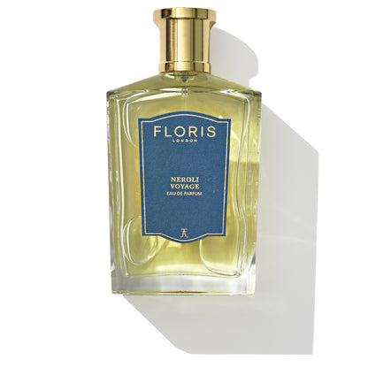 A clear rectangular glass bottle of Floris London Neroli Voyage - Eau de Parfum, a unisex citrus marine fragrance, features a gold cap and a blue label, casting a shadow on a white surface.