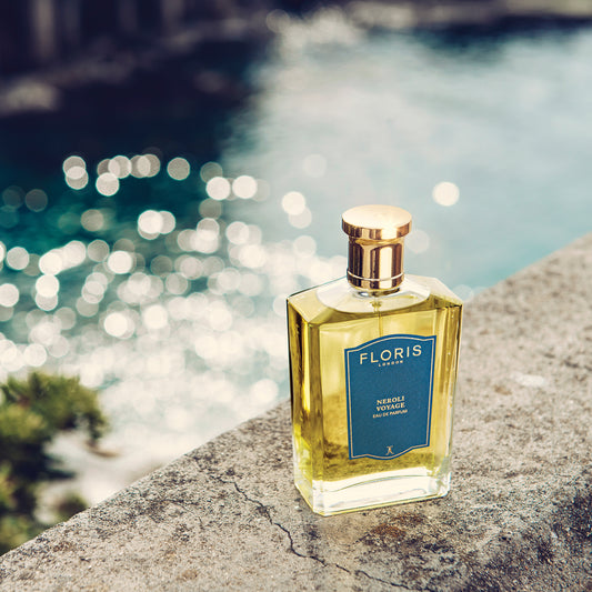 A bottle of Floris London Neroli Voyage - Eau de Parfum rests on a stone ledge, evoking a sunlit citrus-marine scent by blue waters.