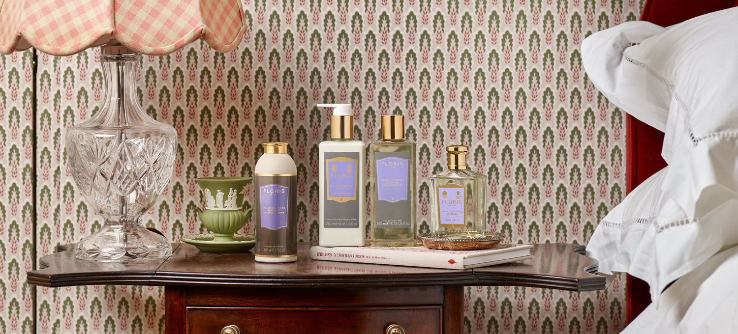 The Night Scented Jasmine collection sits on a stack of books atop a wooden nightstand against a backdrop of floral wallpaper.