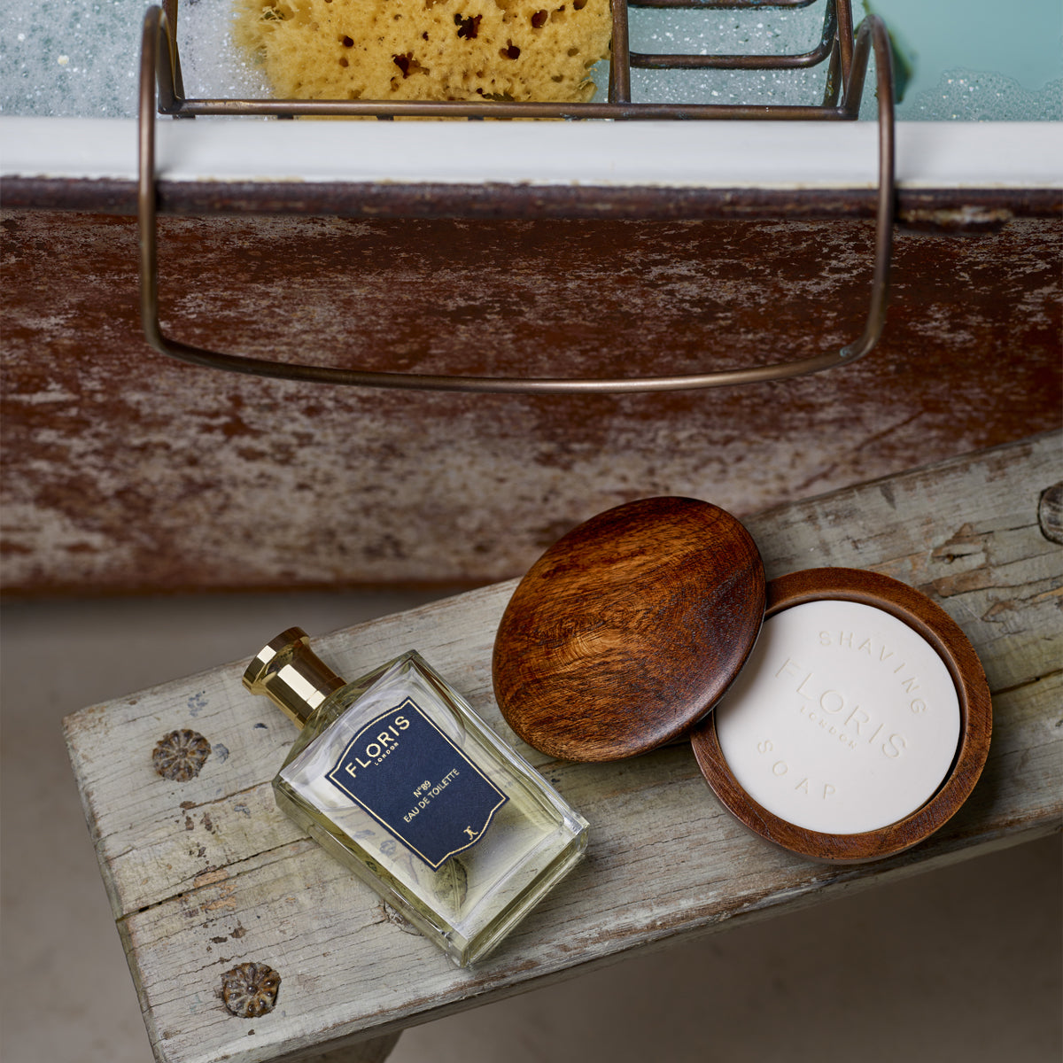 A tube of Floris London No. 89 Shaving Cream sits on a rustic wooden bench by the bathtub, next to a shaving soap dish and sponge.
