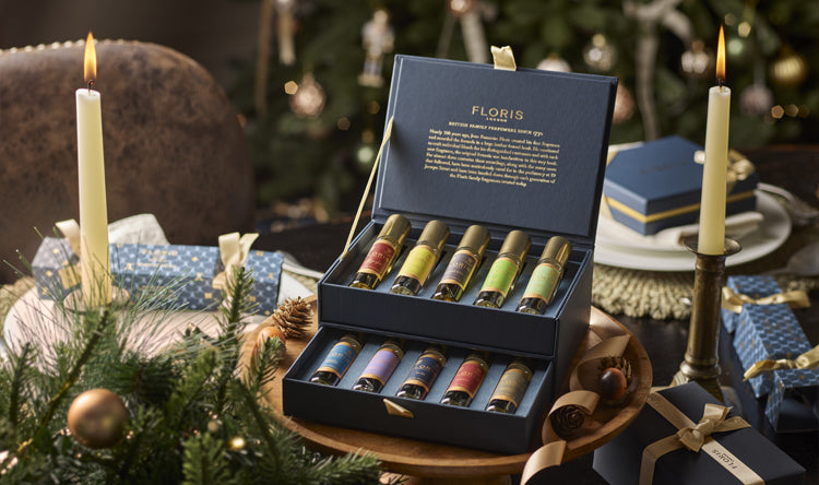 A box of assorted perfumes from The Perfumer's Collection by Floris London, with festive decor in the background, featuring a lit candle and wrapped gifts, makes for the perfect gift for any fragrance connoisseur.